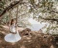 Young European beautiful girl in white bridal marriage dress posing on swing in forest trees on the ocean sea beach Royalty Free Stock Photo