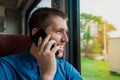 Young european brunette guy in a blue shirt laughs and smiles, looking out the window and talking on a mobile phone in a modern Royalty Free Stock Photo