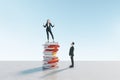 Young european businessman looking up at happy female colleague standing on book pile. Blue sky background. Education, knowledge Royalty Free Stock Photo