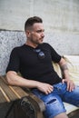 Young European male with a black shirt and shorts sitting near a concrete building at daytime Royalty Free Stock Photo