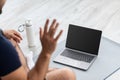 Young european male sits on mat on floor and waves his hand on laptop with empty screen in room