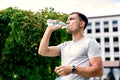 Young European man drinking water from plastic bottle on background of green bushes of trees on bright Sunny day Royalty Free Stock Photo