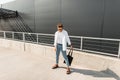 Young European man hipster in a white shirt in blue fashionable jeans in dark sunglasses with a black bag posing in the street Royalty Free Stock Photo