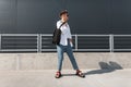 Young European man hipster in a white shirt in blue stylish jeans in dark sunglasses with a black bag stands in the city Royalty Free Stock Photo