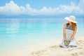 Young european woman in dress, hat and with green apple is sitting on the sand beach of calm tropical sea at sunny day