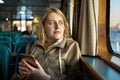 Young European woman inside passenger cabin of ferry looks out window and holds smartphone