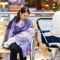 Young european woman with violet stole is breastfeeding her little child close to white baby carriage at public place shopping mal