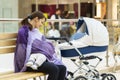 Young european woman with violet stole is breastfeeding her little child close to white baby carriage at public place shopping mal