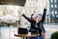 Young excited cheerful brunette girl in leather jacket, at outdoor city cafe, sitting with arms raised after playing Royalty Free Stock Photo