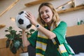 young excited female football fan in green t-shirt and scarf holding ball and celebrating during watch of soccer match Royalty Free Stock Photo