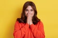 Young excited happy surprised wondered beautiful woman wearing casual orange sweater, covering mouth with hand, looking at camera