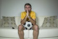 Young excited and nervous football supporter man watching soccer game on television at living room sofa feeling emotion and stress