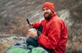 Young explorer bearded man drinking tea or coffee in mountains. Traveler man wearing red clothes holding in hands a mug of tea