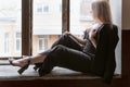 Young fair-haired woman in black pantsuit and heels sits on the windowsill of an old building and looks out the window