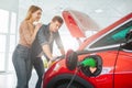 Young family buying first electric car in the showroom. Attractive couple looking under eco-friendly car hood. Electric