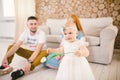 Young family sits on the floor near the couch,small daughter of a blonde one year old is learning to walk in a white dress against Royalty Free Stock Photo