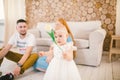 Young family sits on the floor near the couch,small daughter of a blonde one year old is learning to walk in a white dress against Royalty Free Stock Photo