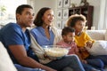 Young family sitting together on the sofa in their living room watching TV and eating popcorn, side view