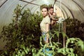 Young family of three in greenhouse, woman is watering tomatoes, man is holding six month old baby daughter in his arms.