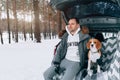 A young family of travelers a guy are sitting in the trunk of their car in an embrace with their friend dog Beagle. Winter forest