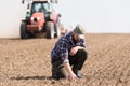 Young farmer examing dirt while tractor is plowing field Royalty Free Stock Photo