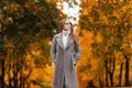 Young fashionable hipster woman in elegant long coat with glasses in a knitted white sweater stands in the park on a warm autumn Royalty Free Stock Photo