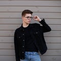 Young fashionable man hipster straightens stylish glasses near wooden wall in street. Handsome guy model in youth black denim