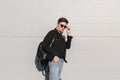 Young fashionable man hipster straightens stylish sunglasses near vintage wall on bright sunny day. Handsome guy model in black Royalty Free Stock Photo