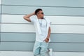 Young fashionable man hipster in youth stylish summer white and jeans clothes in dark sunglasses black rests outdoors near a Royalty Free Stock Photo