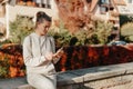 Young Fashionable Teenage Girl With Smartphone In Europian Park In Autumn Sitting At Smiling. Trendy Young Woman In Fall Royalty Free Stock Photo