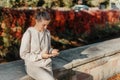 Young Fashionable Teenage Girl With Smartphone In Europian Park In Autumn Sitting At Smiling. Trendy Young Woman In Fall Royalty Free Stock Photo