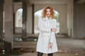 Young fashionably dressed red-haired girl with curly hair in a white coat posing, looking at the camera in an abandoned room