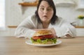 Young fat excited woman sitting at the table in kitchen looking at the plate with big burger.