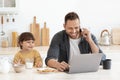 Young father businessman working on laptop and talking with client on phone, cute son enjoying healthy snack at kitchen Royalty Free Stock Photo