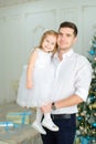 Young father keeping little daughter wearing white dress near Christmas tree.