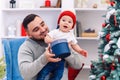 Young father sits in comfortable chair holding amusing toddler and gives gift box to him in the wonderfully decorated