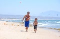 Young father and son running along beach with surfboard