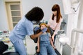 Young female African American dentist with dental tools, making dental check up of her little patient girl. Young mother