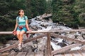 Young female backpacker resting on the mountain cold water stream bridge. Woman sitting on bridge fence smiling and enjoying the