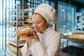 Young female baker smelling fresh bread over modern bakery background