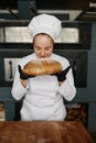 Young female baker smelling fresh bread over modern bakery background