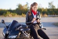 Young female biker with phone in hands, typing message on cellular, wears red bandana, white shirt and leather jacket, poses on