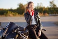 Young female biker with serious expression, has phone conversation via cellular, wears red bandana and leather jacket, poses on mo