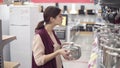 Young female costumer choosing cook pot from a variety of kitchenware in an appliance store. Examining carefuly steel