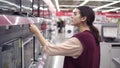 Young female costumer choosing new TV set compares pricetags for different TV screens in department of electronics in