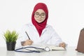 Doctor with hijab, sitting and writing something in her book over white background Royalty Free Stock Photo