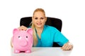Young female doctor or nurse sitting behind the desk and holding piggybank