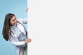 Young female doctor peeking from behind white blank placard board for medical text or design, banner Royalty Free Stock Photo