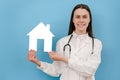 Young female doctor in professional medical white uniform and stethoscope holds small paper house, looking at camera smiling, Royalty Free Stock Photo