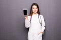 Young female doctor showing mobile phone against grey background